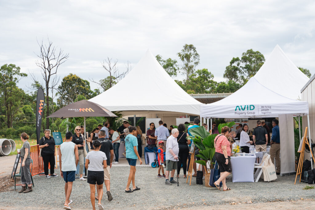 A group of people gathers outside under white tents at an outdoor event surrounded by trees. Some people are talking, while others visit stalls with banners and displays. The atmosphere appears casual and social.