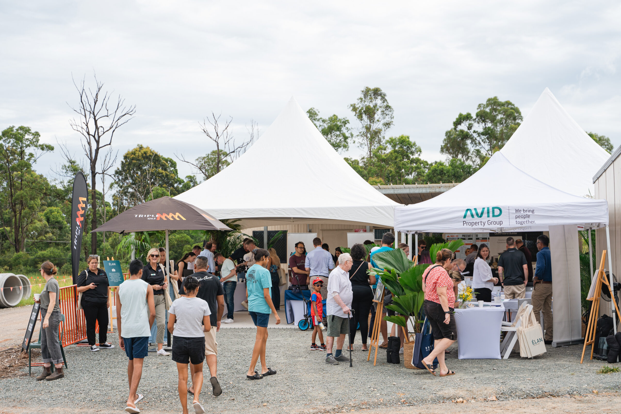A group of people gathers outside under white tents at an outdoor event surrounded by trees. Some people are talking, while others visit stalls with banners and displays. The atmosphere appears casual and social.