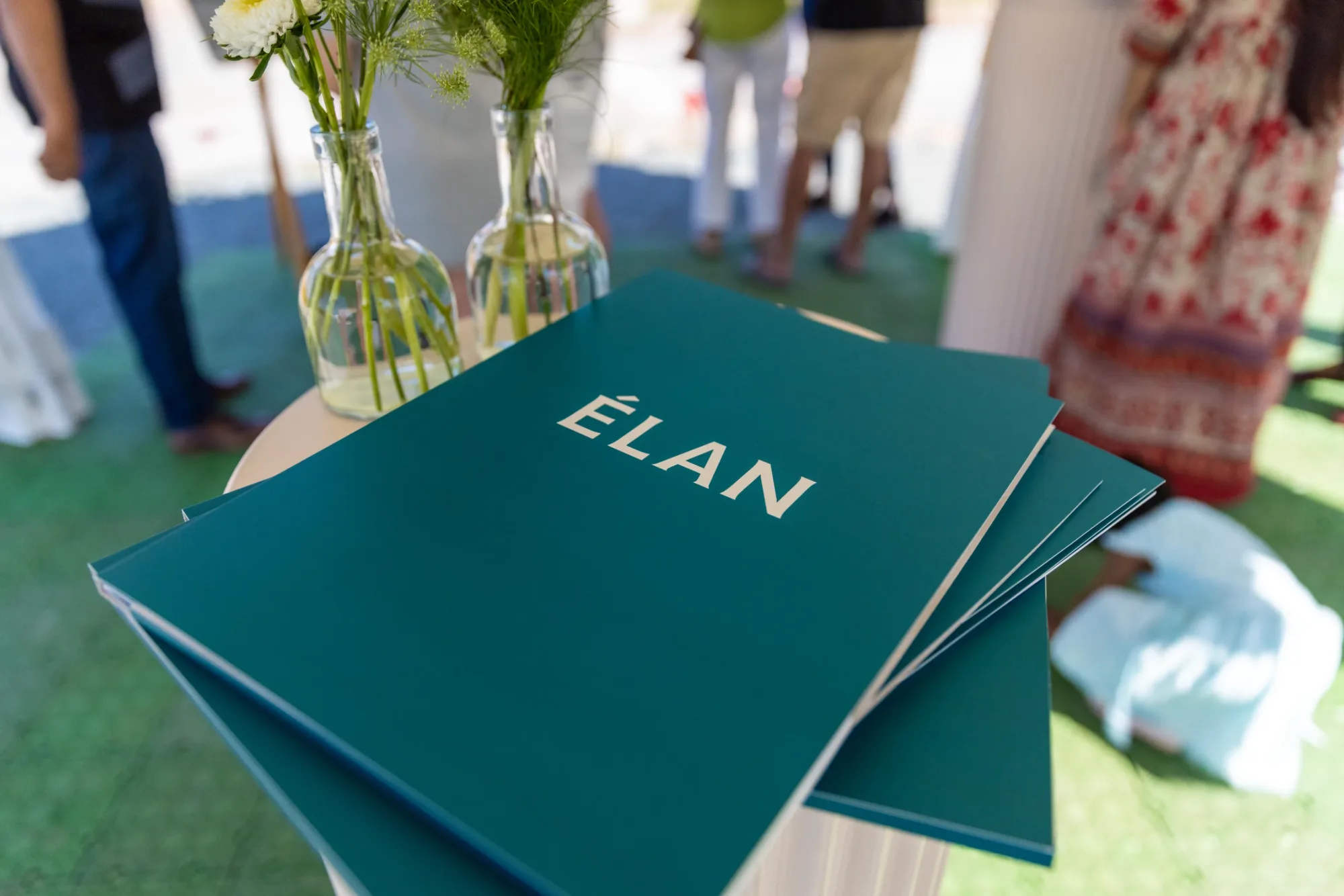 A stack of teal folders labeled “ÉLAN” sits on a small round table next to glass vases with flowers. People stand in the blurred background.