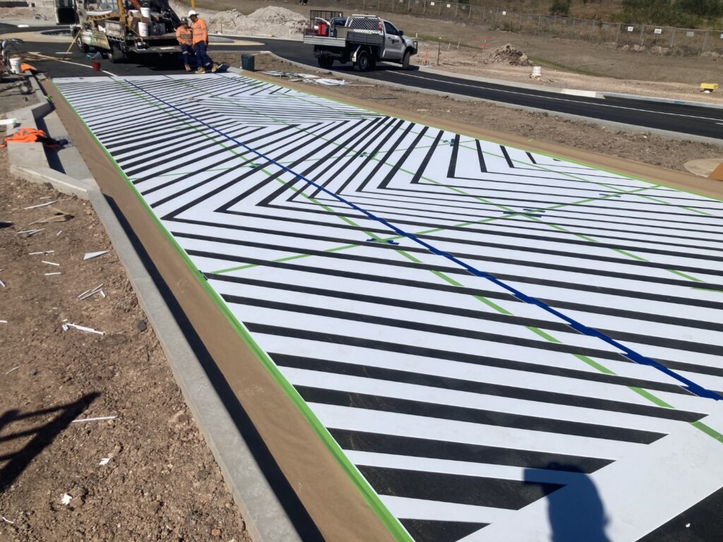 A large geometric mural with black and white diagonal stripes is being installed on the ground. Construction workers and vehicles are visible nearby at the worksite.