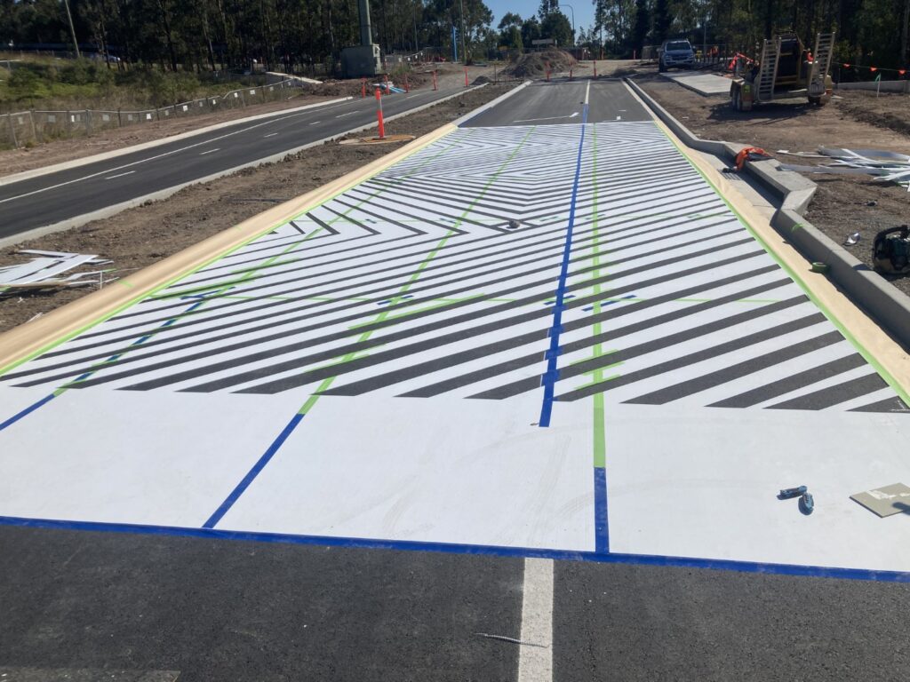 A wide pedestrian crossing with bold black and white diagonal stripes is being constructed on a road. Construction materials and equipment are visible nearby, and the surrounding area is under development.