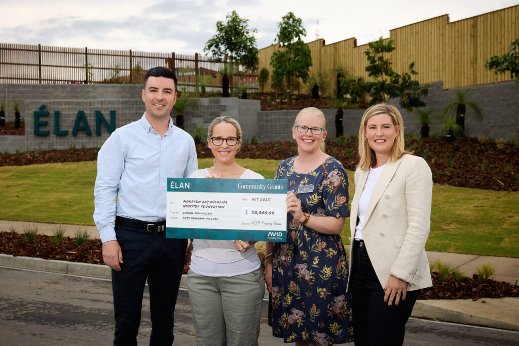 Four people standing outdoors, smiling and holding a large cheque together. The cheque displays a grant amount. There is greenery and a sign reading "ELAN" in the background.
