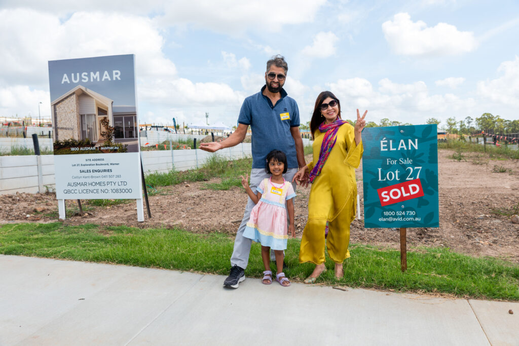 A smiling family of three poses in front of a sold lot sign and a home builder sign at a construction site. The parents and their child stand on a sidewalk with empty land behind them.