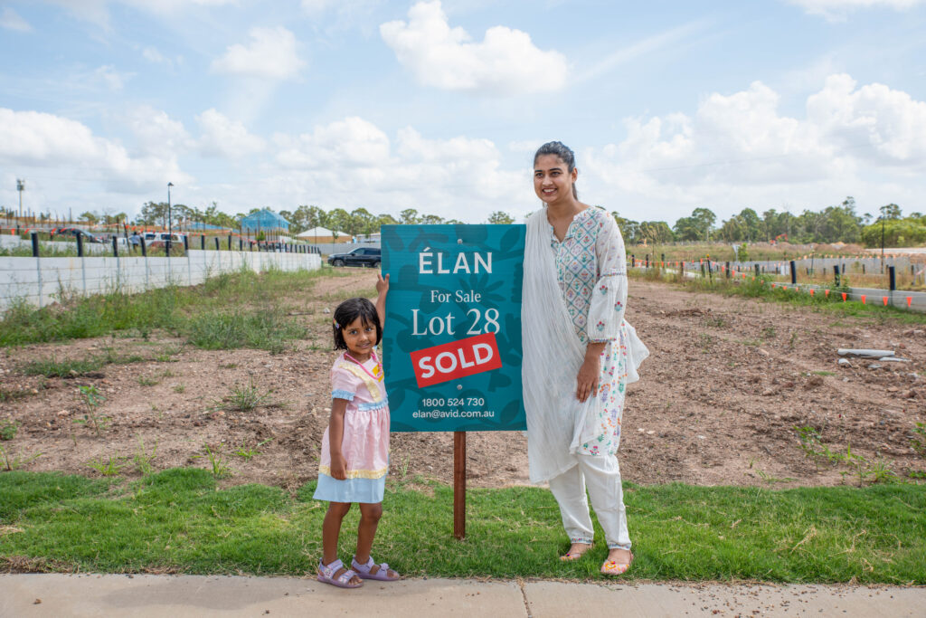A woman and a young girl stand smiling beside a "Lot 28 For Sale: SOLD" sign on a vacant plot of land in a suburban area, with trees and houses in the background.