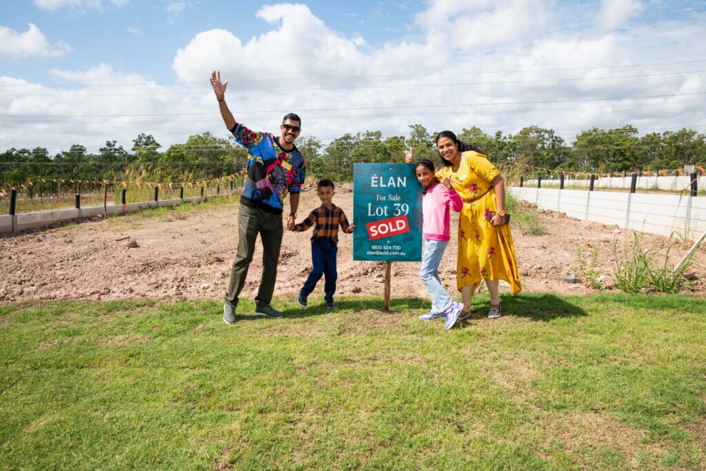 A smiling family of four poses next to a "For Sale" sign that reads "Lot 39 SOLD" on a grassy plot, celebrating their new property on a sunny day.
