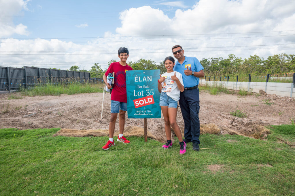 Three people stand smiling next to a sign that reads "ÉLAN For Sale Lot 35 SOLD" on a grassy plot of land, with a blue sky and trees in the background.