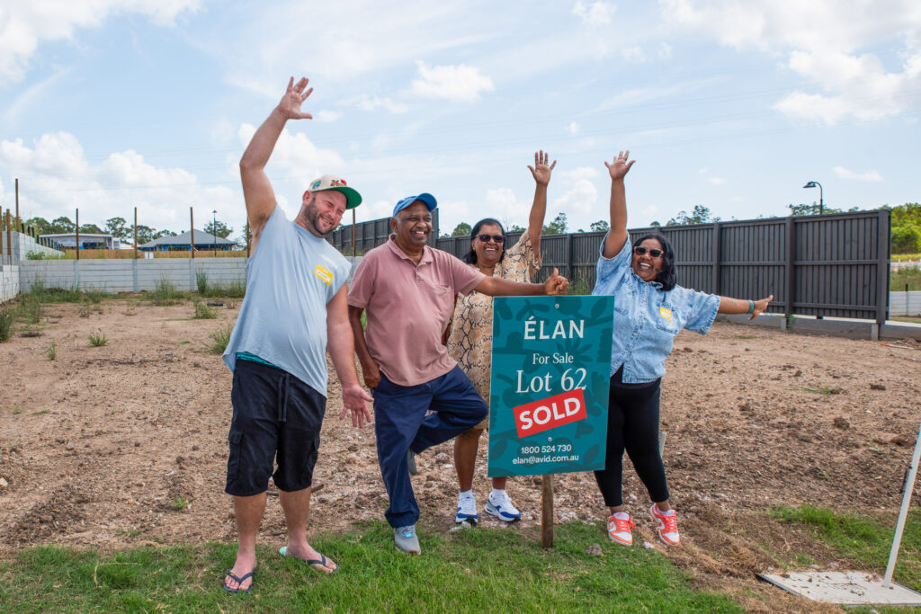 Four people stand smiling and raising their arms in celebration next to a “For Sale” sign marked “Sold” on a dirt plot of land under a partly cloudy sky.