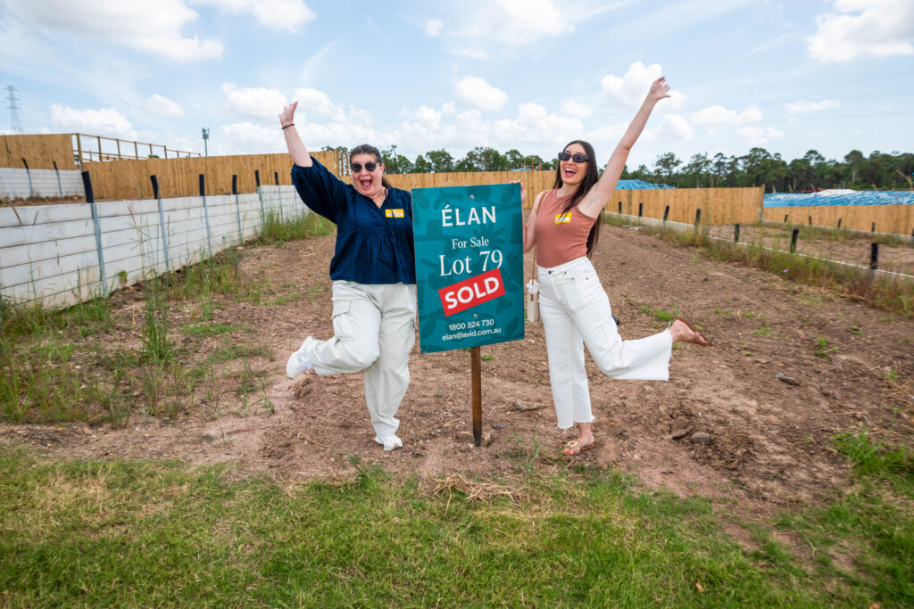 Two women joyfully pose next to a sign that reads "Élan For Sale Lot 79 SOLD" on a dirt plot of land, celebrating the purchase of a new property.