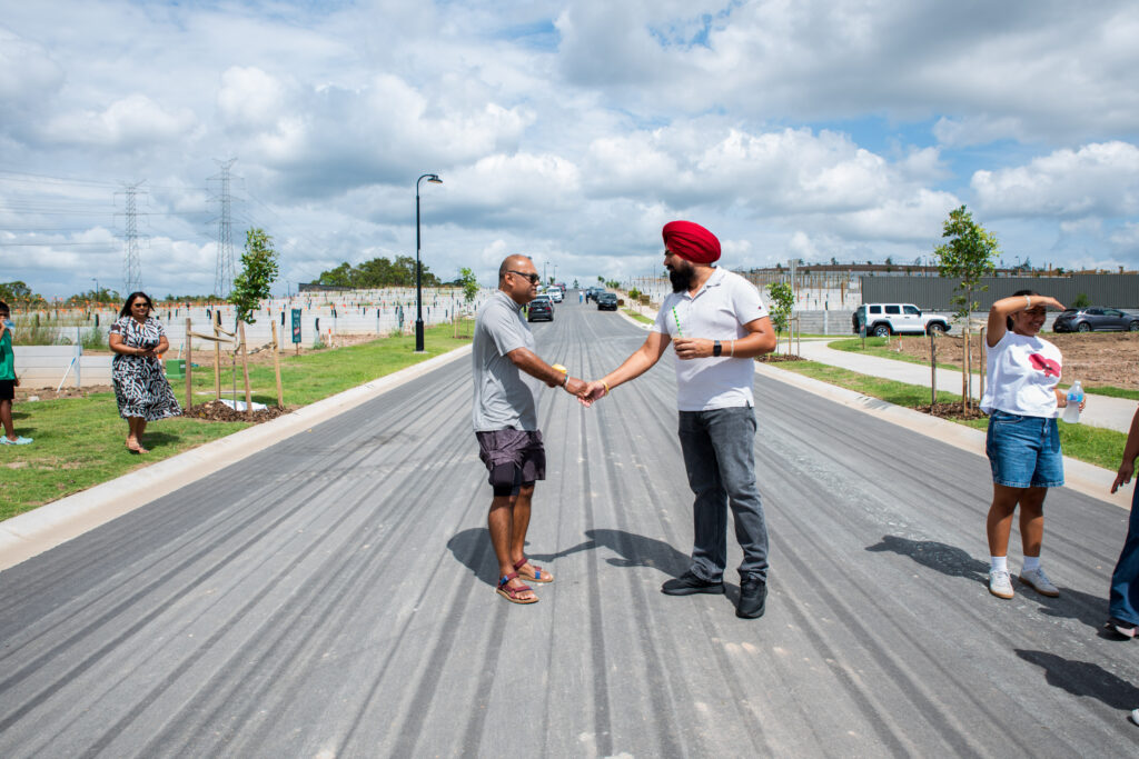 Two men shake hands in the middle of a newly paved street, surrounded by people, trees, and new houses under a partly cloudy sky. One man wears a red turban, the other is in shorts and sandals.