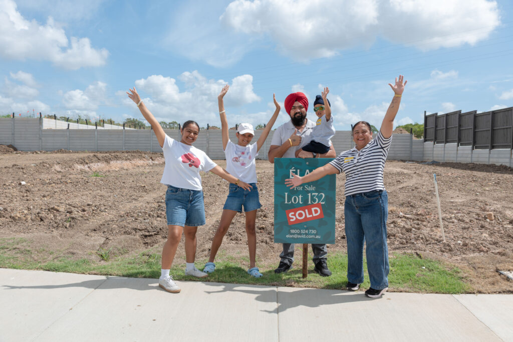 A joyful family of five poses with arms raised next to a “Lot 132 SOLD” sign on a dirt lot, celebrating the purchase of new land on a sunny day.