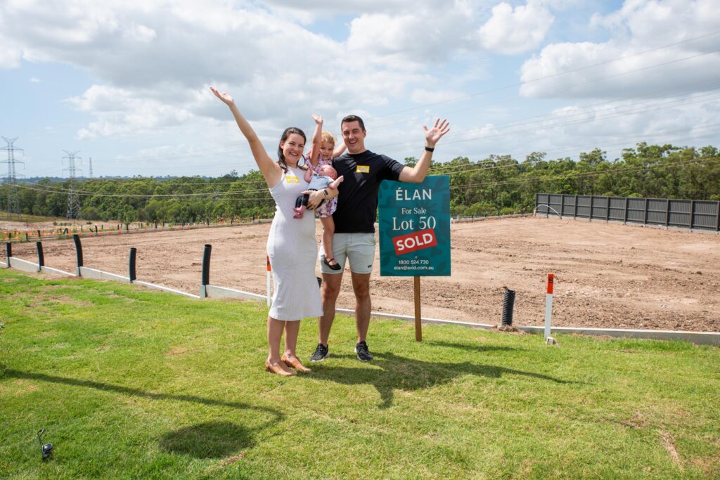 A smiling couple holding a baby stand on grass beside a "Lot 50 Sold" real estate sign, raising their arms in celebration. Behind them is an empty dirt lot, trees, and a partly cloudy sky.