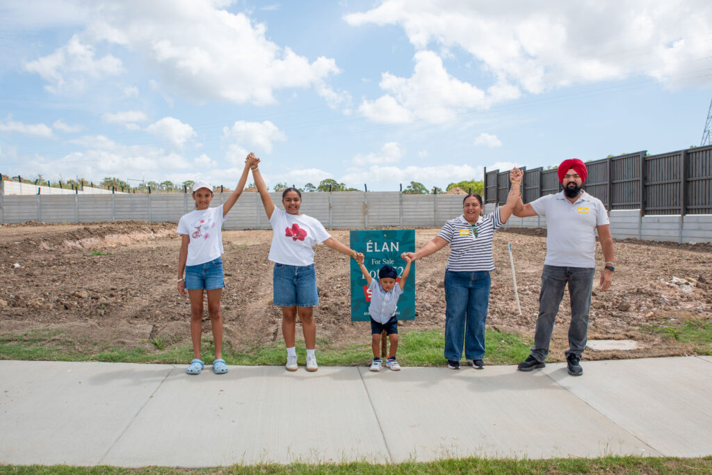 Five people stand hand-in-hand in front of a dirt lot with a green “ÉLAN For Sale” sign. They smile at the camera, holding hands and raising their arms. The sky is partly cloudy, and a fence lines the background.