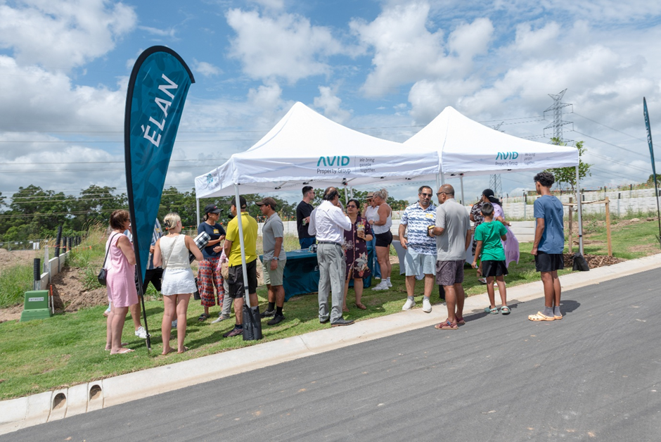 A group of people stands on grass and pavement near a white marquee tent with “AVID” branding, talking and mingling. A blue flag reading “ÉLAN” is displayed beside the tent under a partly cloudy sky.
