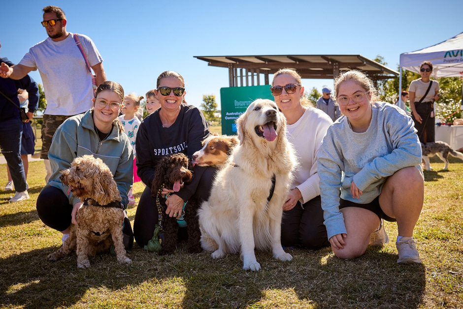 Four smiling women kneel on grass with four happy dogs of various breeds at an outdoor event on a sunny day. Other people and booths are visible in the background.