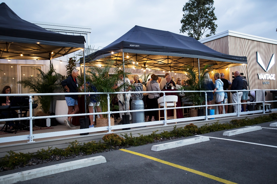 A group of people gathers under black canopies outside a modern building with "VANTAGE" signage. Some are seated, others are standing and talking. Plants decorate the area; it's early evening in a parking lot.
