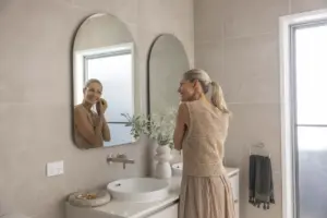 A woman with blonde hair in a ponytail smiles as she looks in the bathroom mirror, putting on an earring. The bathroom is modern with beige tiles, a white sink, and a vase with greenery.
