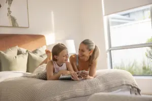 A woman and a young girl are lying on a bed together, smiling and reading a book in a bright, sunlit bedroom with large windows and neutral-colored decor.