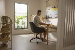 A man sits at a wooden desk in a bright, modern room, working on a laptop. There’s a large window with plants outside, a cup on the desk, and abstract art on the wall.