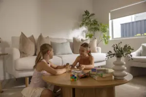 Two young girls sit on the floor in a bright living room, playing a card game at a round wooden coffee table with board games and a Rubik’s cube on top. A white sofa and green plant are in the background.