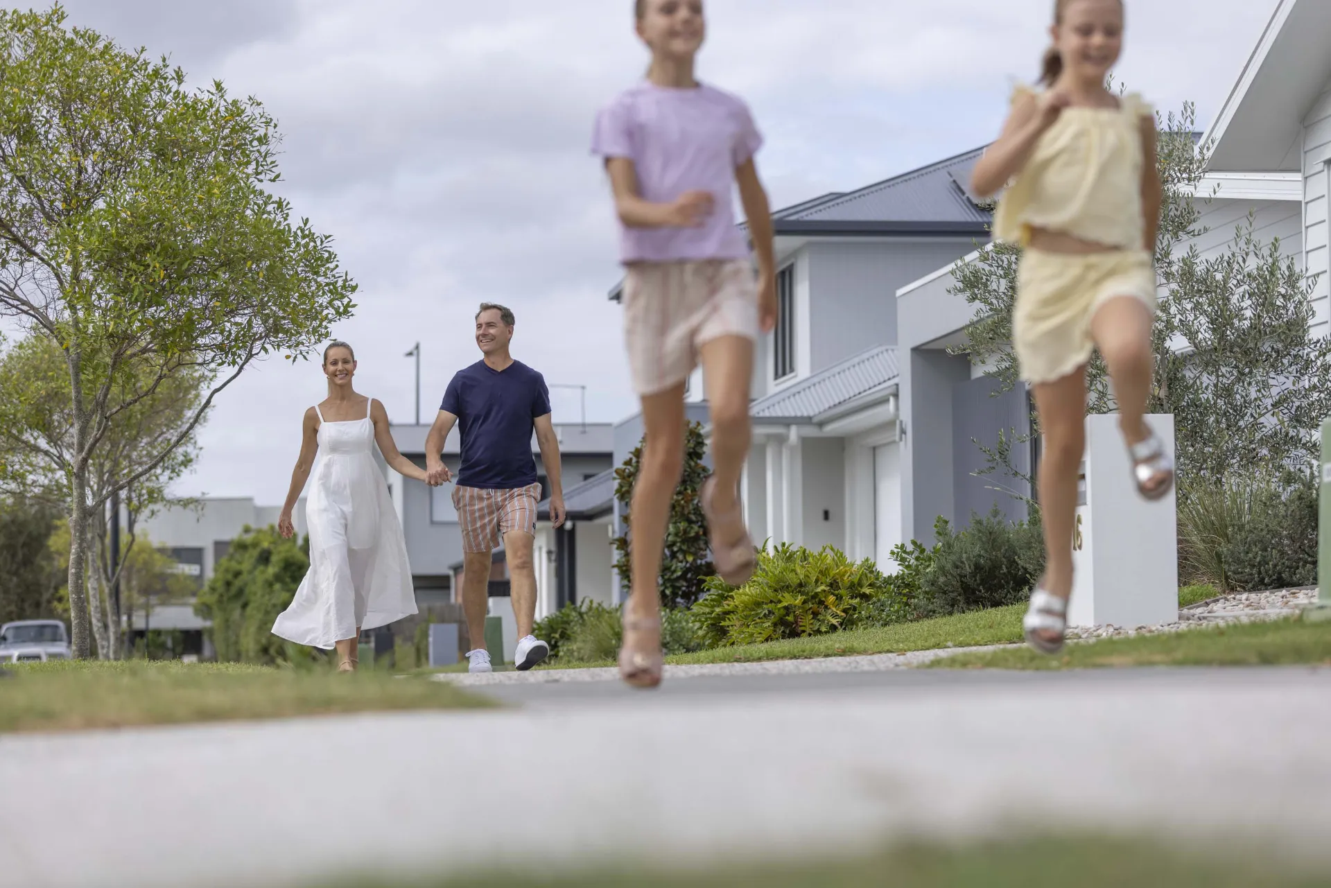 A family walks outside in a suburban neighborhood. Two children run ahead on the sidewalk while two adults walk behind them, holding hands. Trees and houses line the street under a cloudy sky.