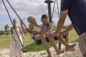 Two young girls laugh together while sitting on a green swing at a playground. An adult woman stands behind them smiling, and a man pushes the swing. The scene is outdoors on a sunny day.