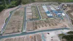Aerial view of a suburban construction site with paved roads, empty lots, and partially built houses; some buildings have blue tarps, and surrounding areas show earthworks and construction equipment.