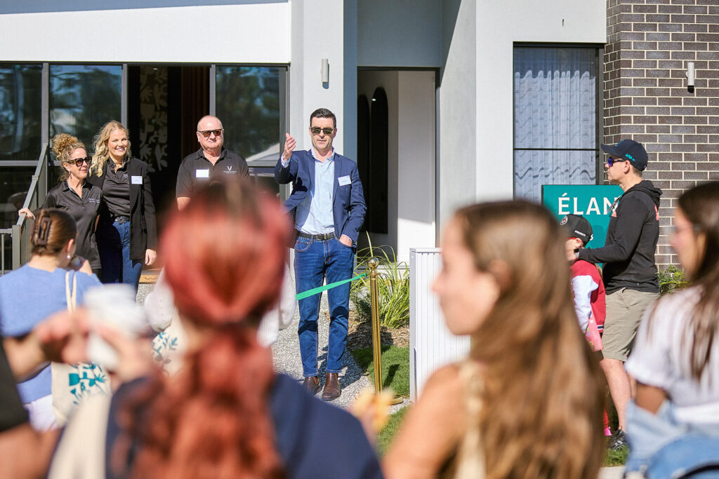 A man in a suit speaks to a crowd in front of a modern building, with three people standing beside him and several people facing him, some holding bags. A green sign with “ÉLAN” is visible on the right.