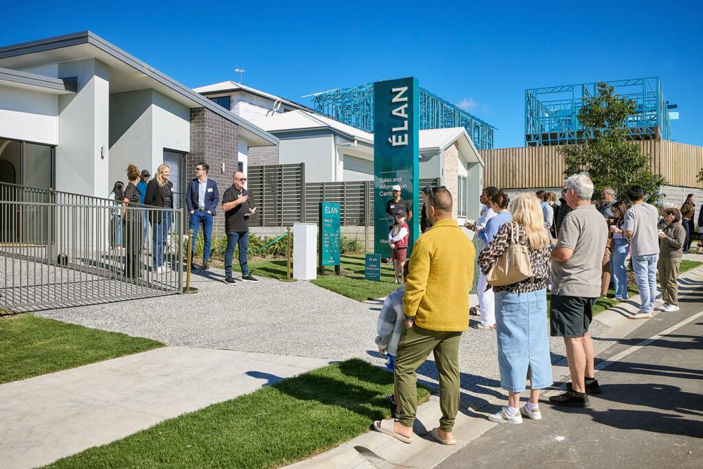 A group of people stand in line outside a modern display home on a sunny day, with signage reading "ÉLAN." Some people wait near the entrance while others walk on the sidewalk.