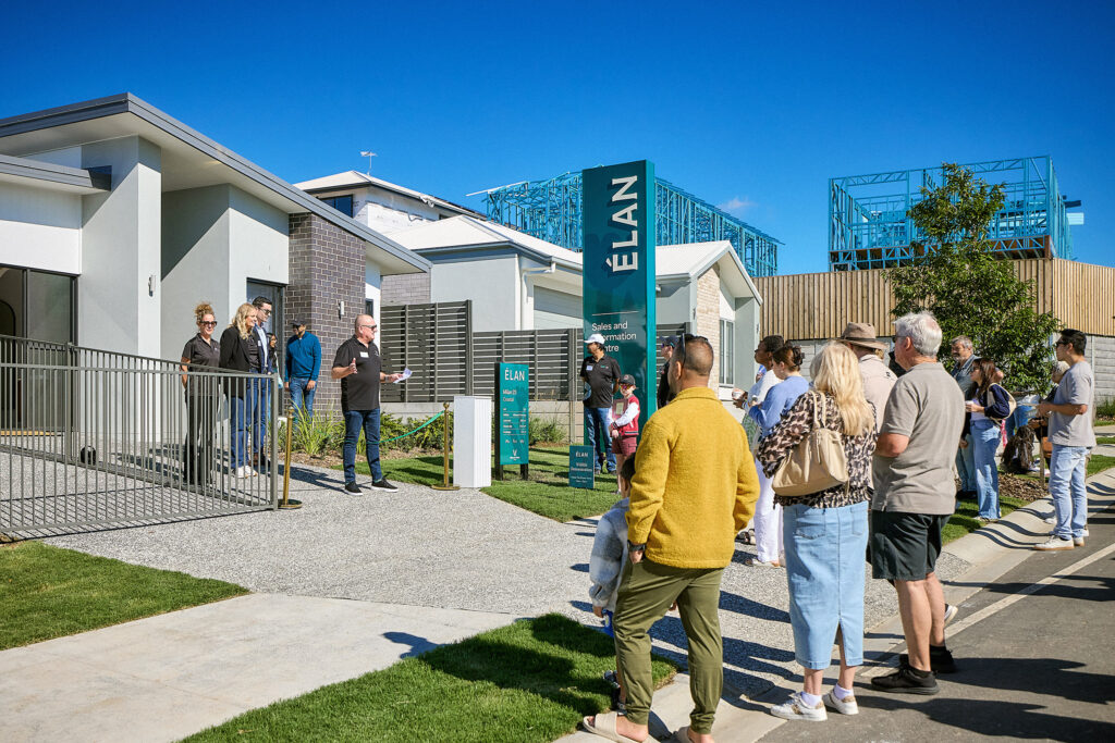 A group of people stands outside a modern house, listening to a person speaking near the entrance. The setting appears to be a real estate open house or auction on a sunny day, with blue skies and other homes visible nearby.