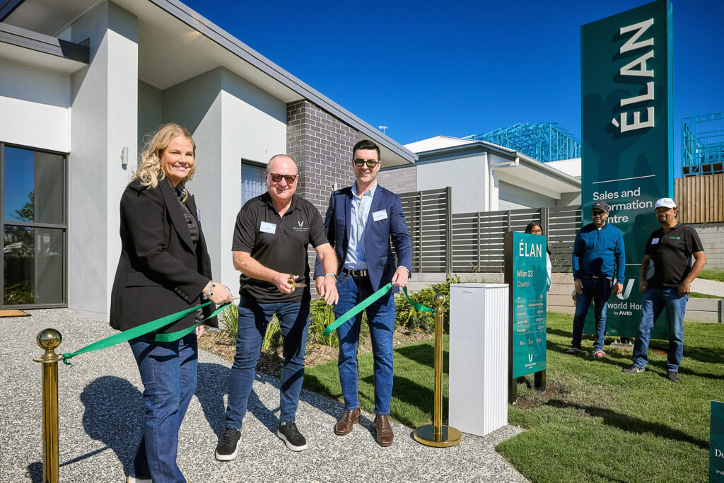 Three people stand in front of a modern building, smiling as they cut a green ribbon at an outdoor ribbon-cutting ceremony for ÉLAN, with a large sign and a few onlookers in the background.