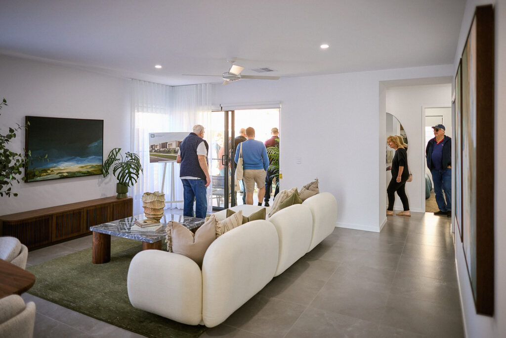 A group of people tour a modern, bright living room with white walls, a plush cream sofa, a green rug, large art pieces, and sliding glass doors leading outside. Some people are entering from a hallway.