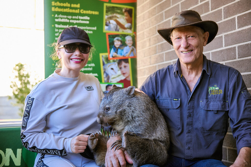 A smiling woman in sunglasses and a man in a hat sit together, holding a wombat. Behind them is a green sign about educational programs. The scene appears to be at a wildlife or educational centre.