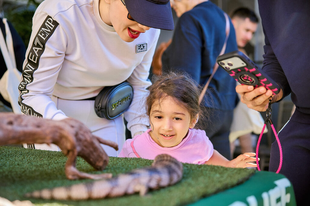 A woman and a young girl smile while looking at a lizard on a green table. Another person holds a phone, possibly taking a photo. The scene appears to be at an outdoor event or exhibit.