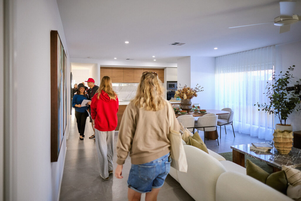 Several people walk through a modern, open-plan living and dining area with large windows, white walls, and stylish decor. The space is bright and airy, with a kitchen visible in the background.