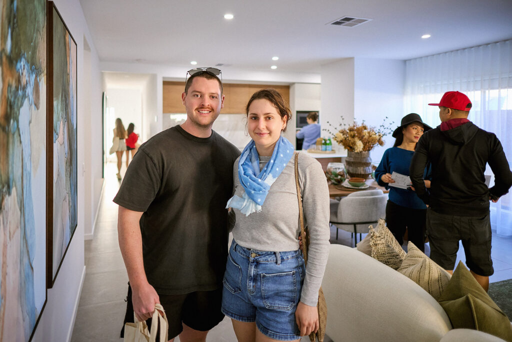 A man and woman smile at the camera in a modern living room. Behind them, three people chat near a dining table. The space is bright, decorated with art, plants, and neutral-colored furnishings.