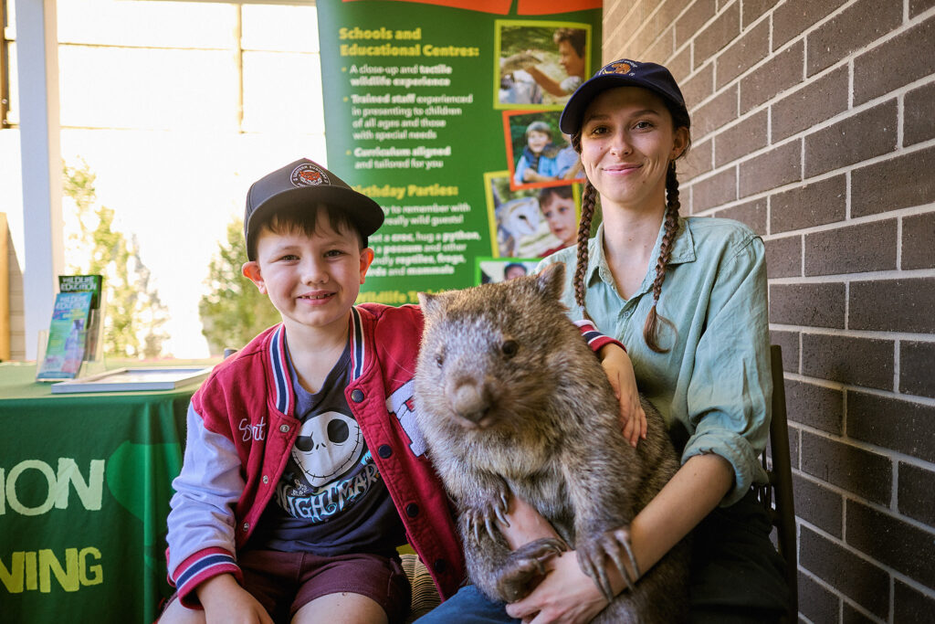A smiling young boy and a woman wearing caps sit together indoors, holding a wombat. Behind them is a green information board about schools and educational centers, and a table with brochures.