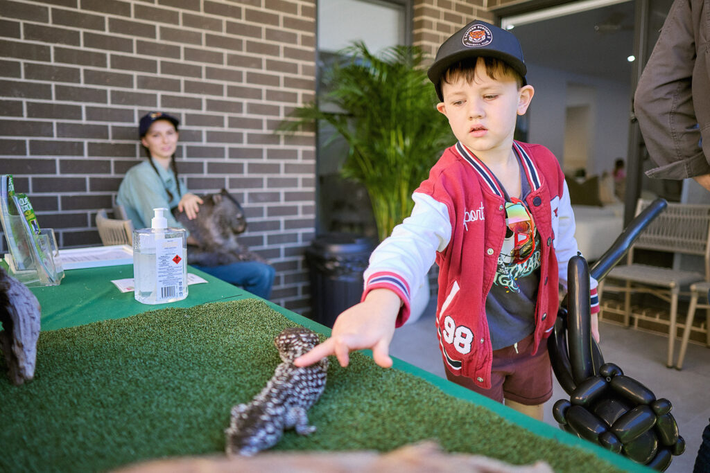 A young boy wearing a red jacket and cap gently touches a lizard on a display table covered with artificial grass. An adult and another person sit in the background near a brick wall.