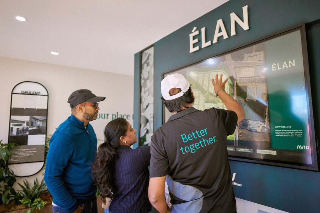 Three people stand in front of a large touchscreen display labeled "ÉLAN" as one person points at a map on the screen; another person wears a shirt that reads "Better together.