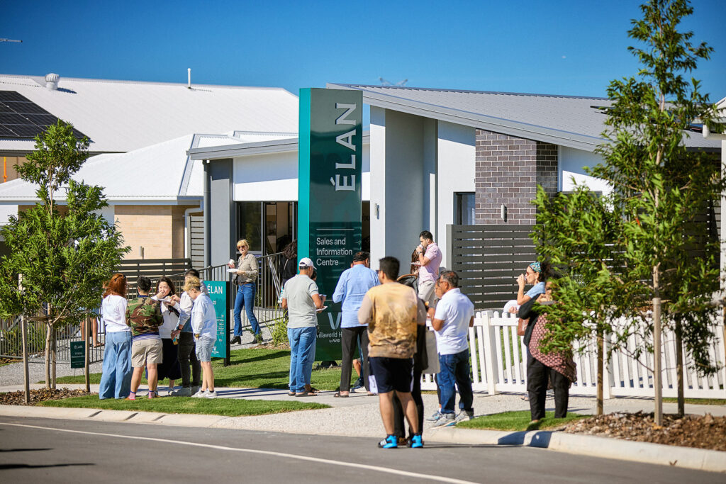 A group of people gather outside the ELAN Sales and Information Centre in a modern residential neighborhood on a sunny day, with houses, greenery, and clear blue sky in the background.