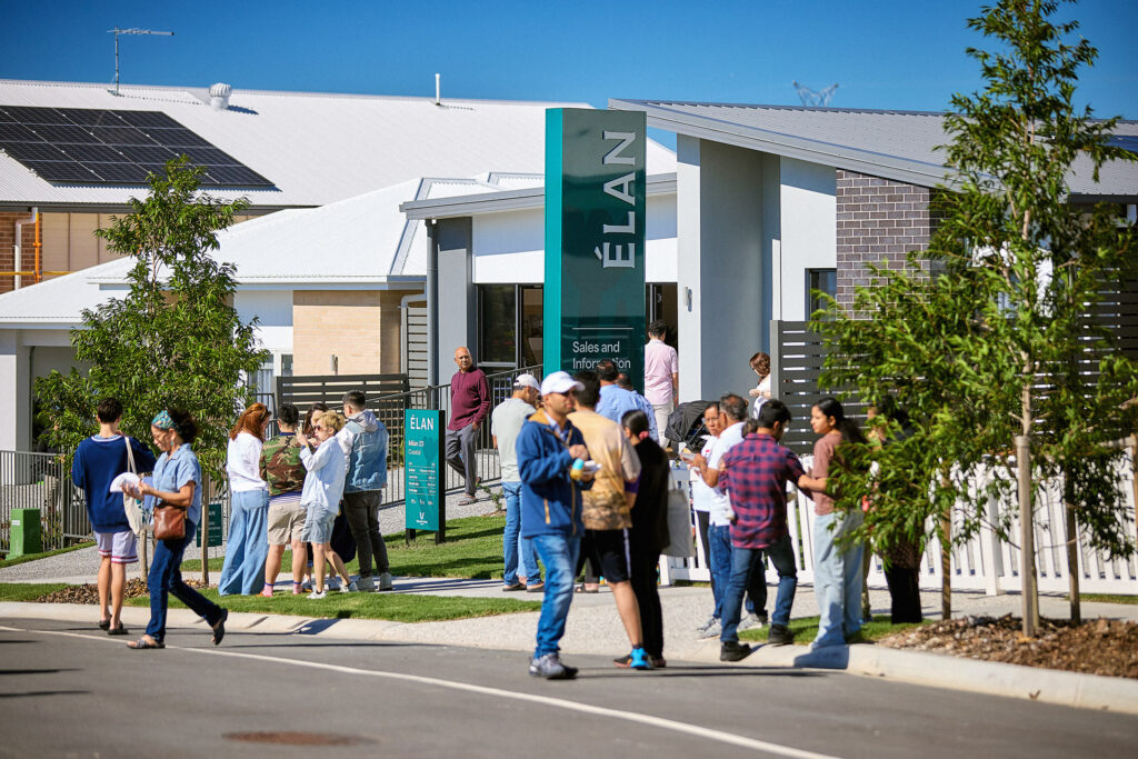 A group of people gather outside a modern residential building with a sign reading "ÉLAN" under a clear blue sky; some people are talking, walking, or holding papers.
