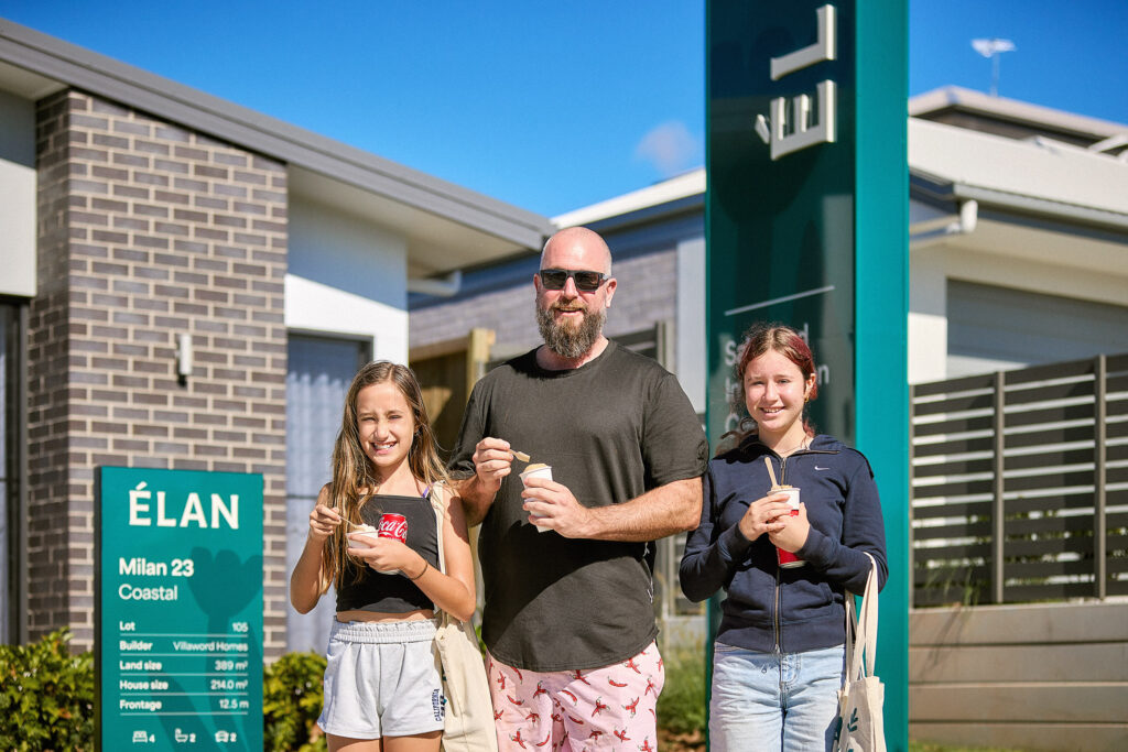 A man and two girls stand smiling outside a modern home display center, holding cups and spoons. A green sign nearby reads “ÉLAN Milan 23 Coastal.” The weather is sunny with a bright blue sky.