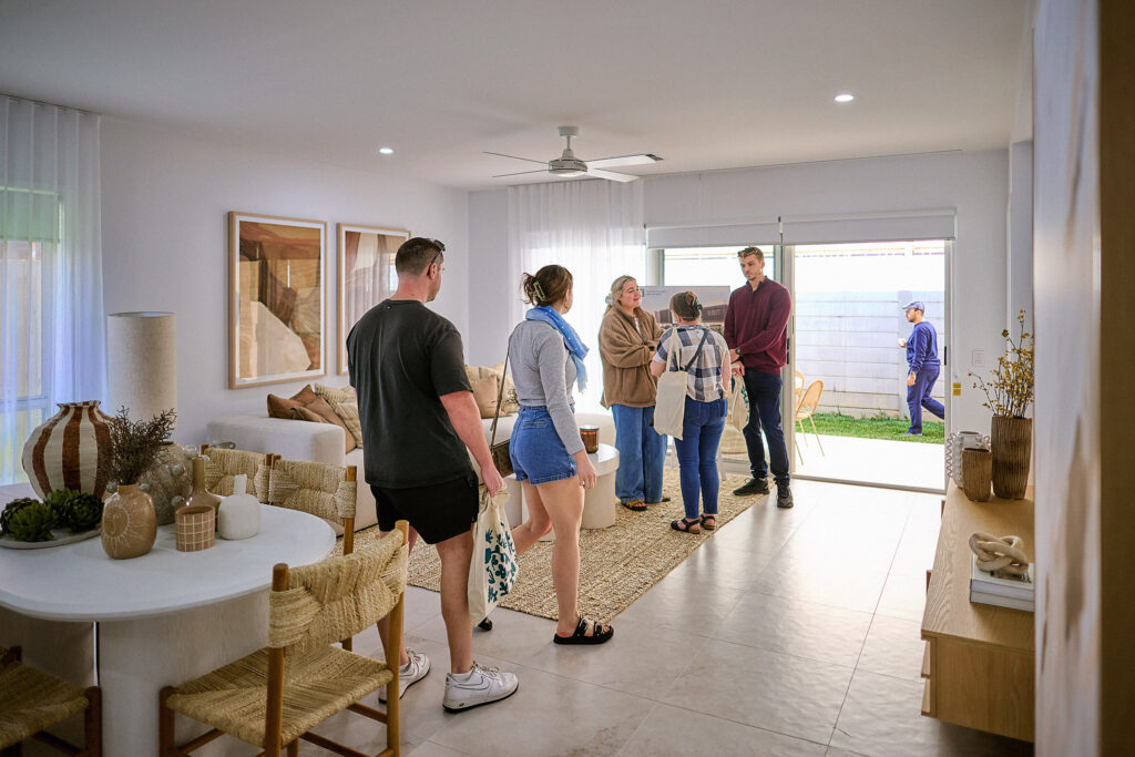 A group of people stand and converse in a bright, modern living room with light-colored decor. Two people are outside on the patio near the glass sliding door, while others gather together inside.