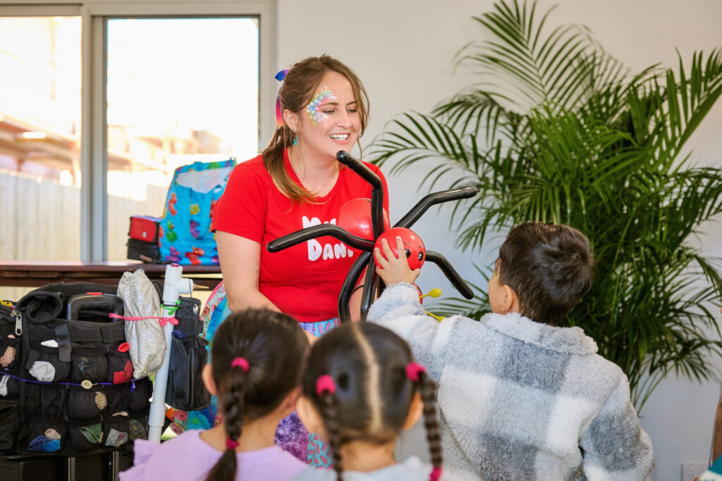 A woman with face paint, wearing a red shirt, smiles and holds a balloon animal in front of three children. One child in a gray coat reaches out to touch the balloon. There are bags and a plant nearby.
