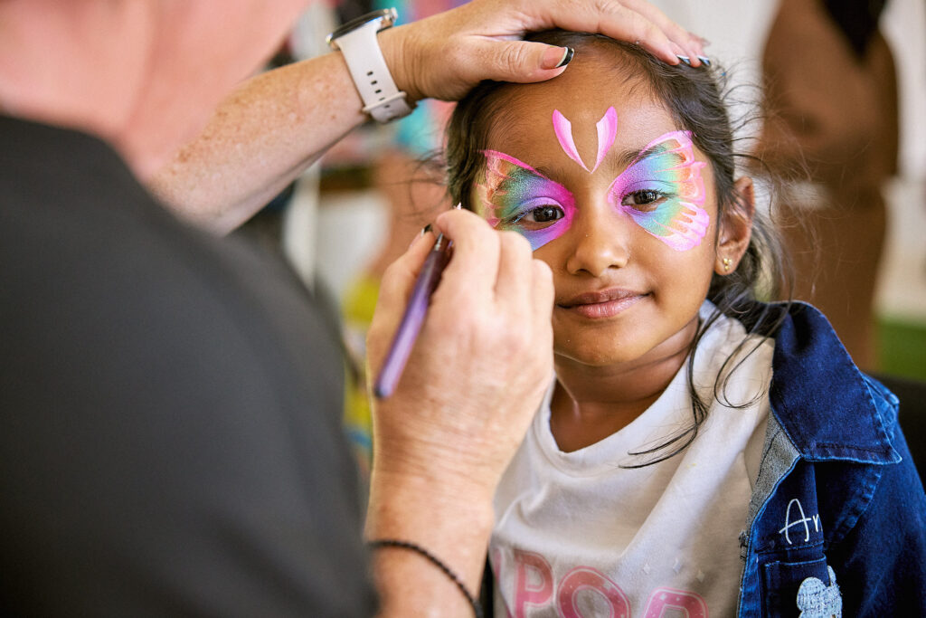 A young girl smiles as an artist paints a colorful butterfly design on her face. The girl wears a denim jacket and a white shirt while the artist carefully applies paint with a brush.