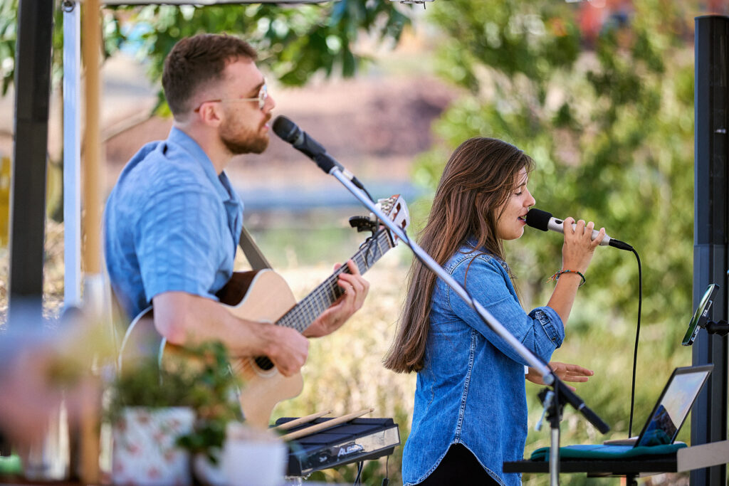 A man playing acoustic guitar and a woman singing into a microphone perform together outdoors under a canopy, surrounded by greenery.