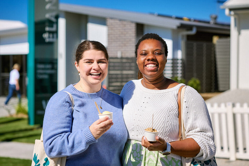 Two women standing outside on a sunny day, smiling and holding cups of ice cream. They are dressed in casual sweaters, and a house with a white fence and a yard is visible in the background.