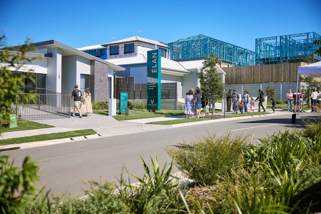 People walk and gather outside modern display homes in a suburban neighborhood under a clear blue sky, with some houses under construction and a green sign reading "ELAN" visible in the center.