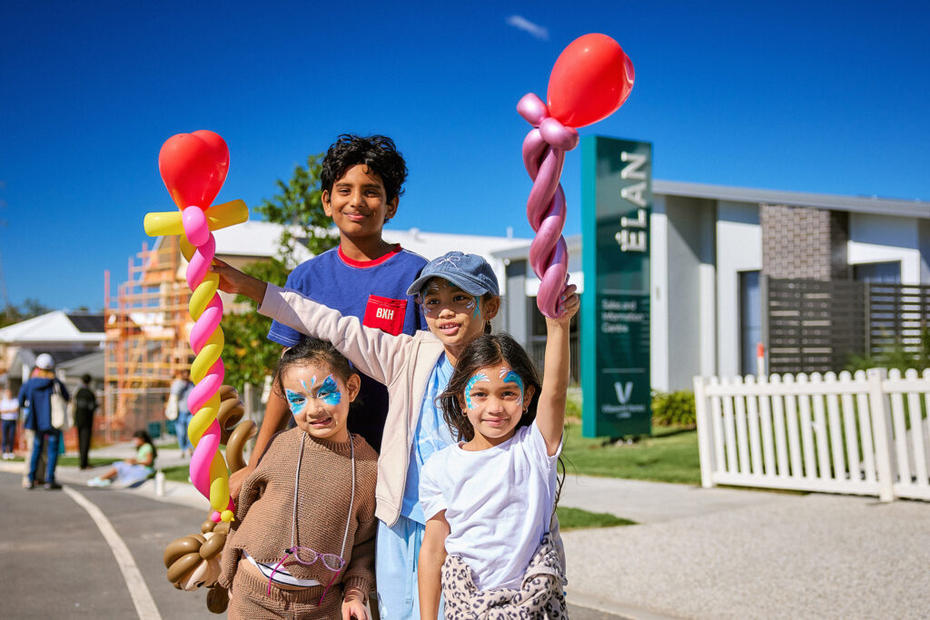 Four smiling children with face paint hold balloon sculptures outdoors on a sunny day, with houses, a white fence, and a construction site visible in the background.