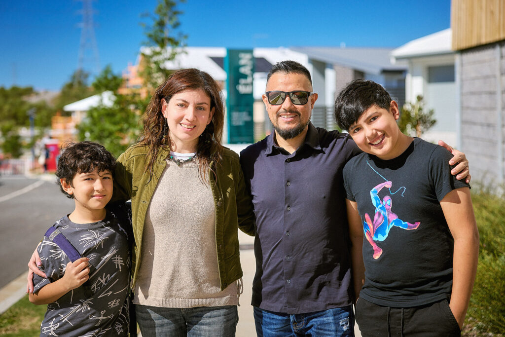 A family of four stands outdoors on a sunny day, smiling at the camera. Two adults are in the center, flanked by two boys. Houses and greenery are visible in the background.