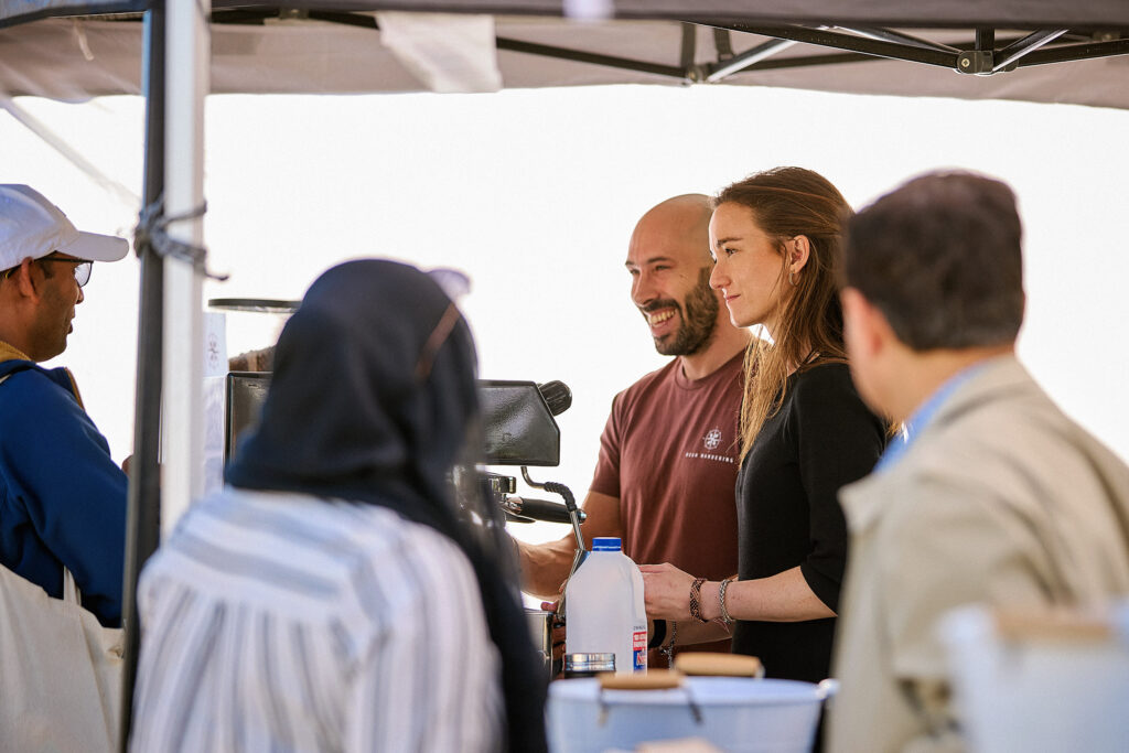 Two people stand behind a coffee counter, smiling and talking to customers. There are bottles and a coffee machine on the counter, with four people gathered around under a canopy.